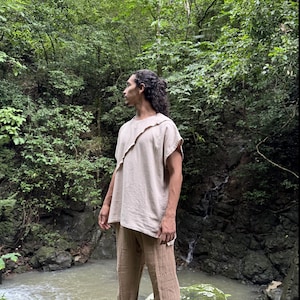 May include: A beige linen top with a unique asymmetrical neckline and matching beige linen pants. The outfit is loose-fitting and appears to be made of a lightweight fabric. The person is standing in a lush, green forest setting.