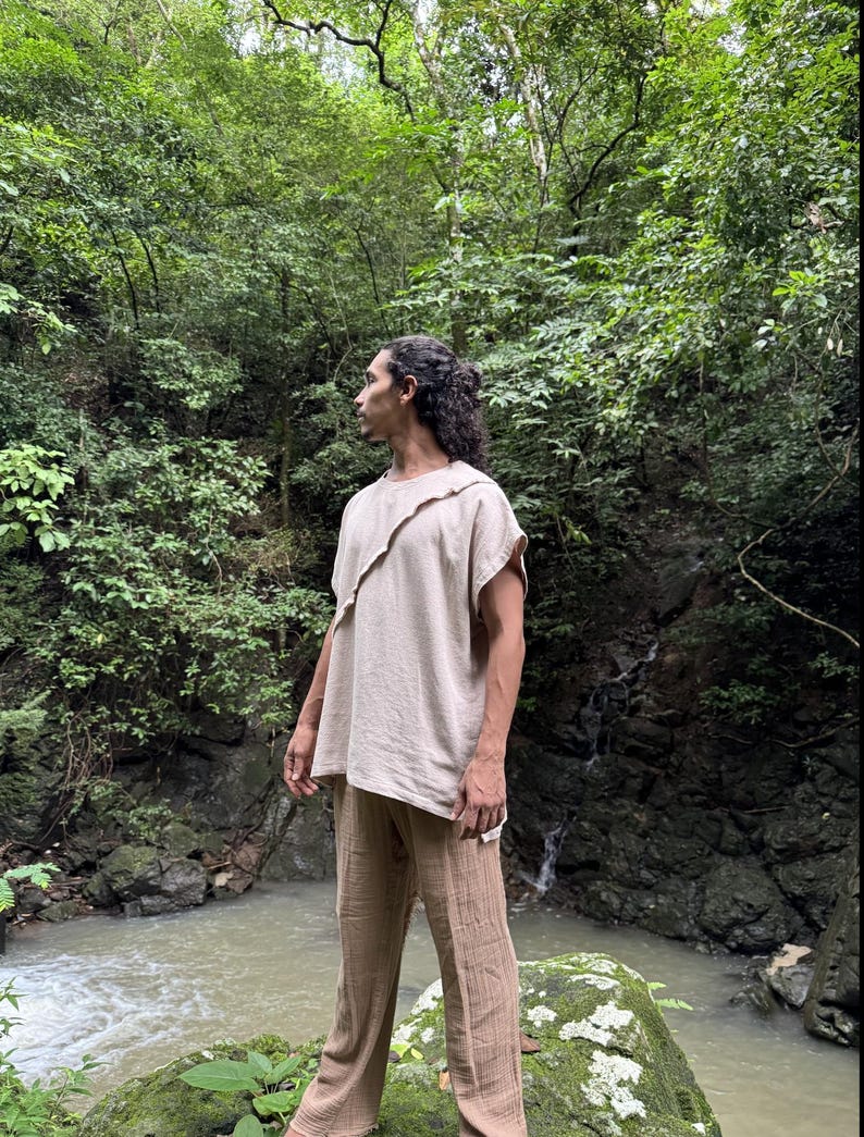 May include: A beige linen top with a unique asymmetrical neckline and matching beige linen pants. The outfit is loose-fitting and appears to be made of a lightweight fabric. The person is standing in a lush, green forest setting.