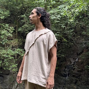 May include: A beige, short-sleeved top with an asymmetrical design. The top has a unique, layered neckline and is paired with matching beige pants. The outfit is made of a textured fabric, suggesting a natural fiber. The person is standing in a lush, green environment.