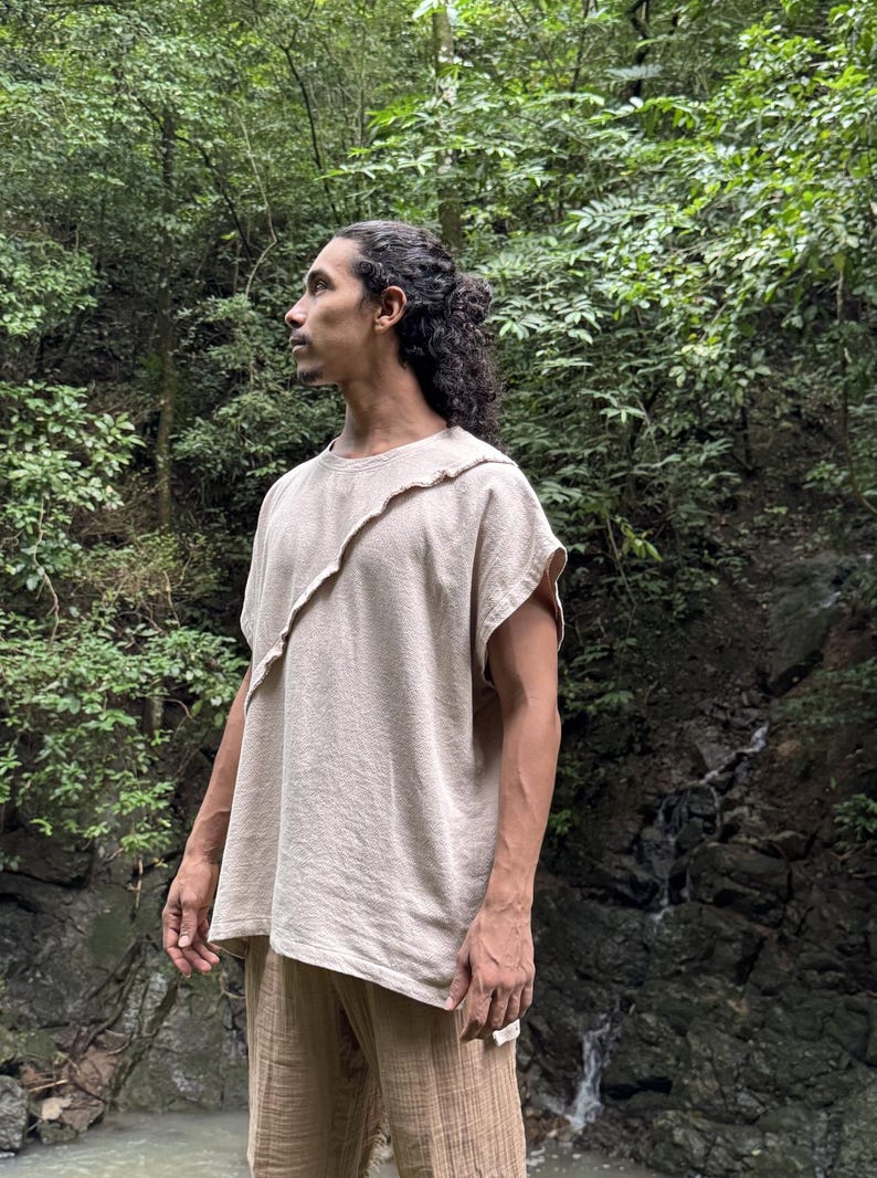 May include: A beige, short-sleeved top with an asymmetrical design. The top has a unique, layered neckline and is paired with matching beige pants. The outfit is made of a textured fabric, suggesting a natural fiber. The person is standing in a lush, green environment.