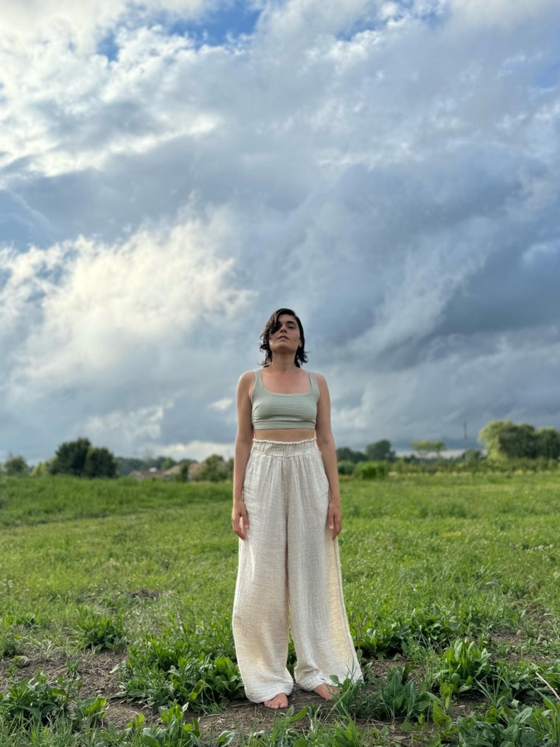 K&ouml;nnte beinhalten: Eine Person tr&auml;gt ein hellgr&uuml;nes Tanktop und eine wei&szlig;e, weite Hose und steht auf einem Feld mit gr&uuml;nem Gras. Der Himmel ist bew&ouml;lkt und blau.