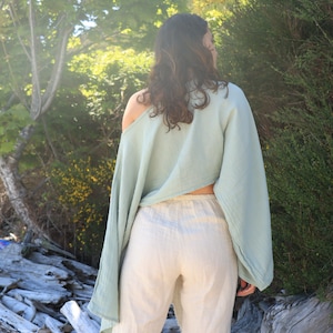 May include: A person wearing a light blue, off-the-shoulder top with long sleeves and white linen pants. The person is standing in a natural setting with trees and driftwood in the background.