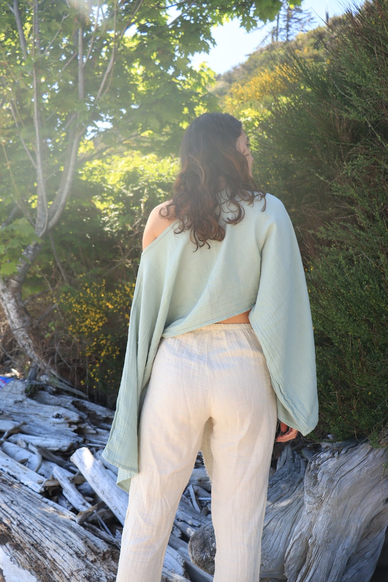 May include: A person wearing a light blue, off-the-shoulder top with long sleeves and white linen pants. The person is standing in a natural setting with trees and driftwood in the background.