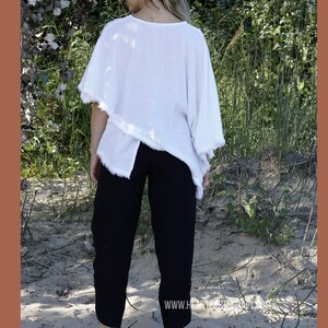 May include: A white, loose-fitting, asymmetrical top with frayed edges, paired with black pants. The model is standing on a sandy beach.