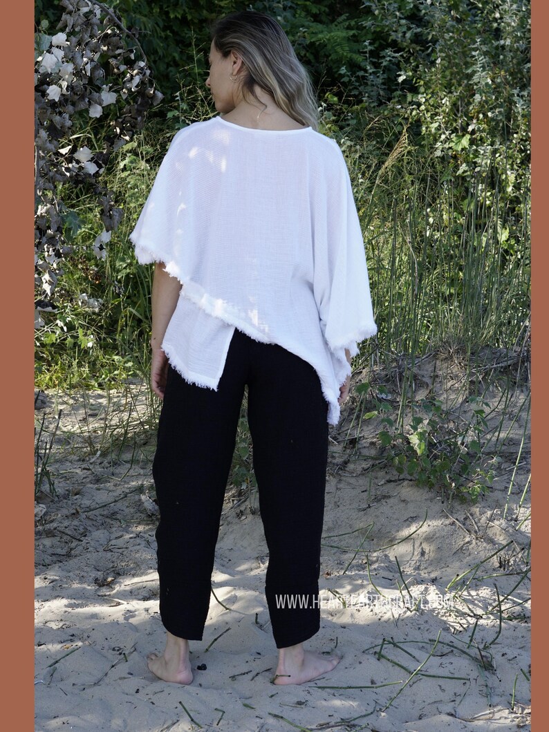 May include: A white, loose-fitting, asymmetrical top with frayed edges, paired with black pants. The model is standing on a sandy beach.