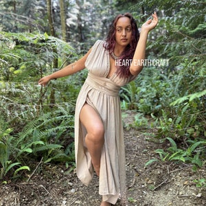 May include: A woman wearing a light brown maxi dress with a deep V-neckline and a slit up the side. She is standing in a forest with green foliage and trees. The dress has a belt at the waist and a beaded necklace. The text "HEART EARTH CRAFT" is visible on the dress.