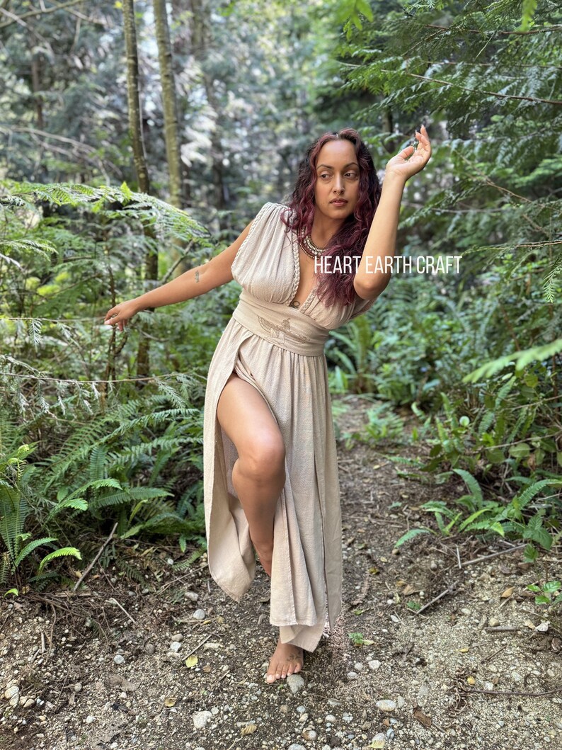 May include: A woman wearing a light brown maxi dress with a deep V-neckline and a slit up the side. She is standing in a forest with green foliage and trees. The dress has a belt at the waist and a beaded necklace. The text "HEART EARTH CRAFT" is visible on the dress.