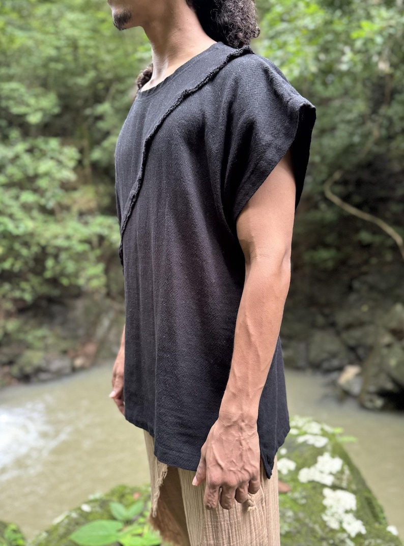 May include: Black linen top with a unique asymmetrical design. The top features a raw edge detail along the neckline and a relaxed fit. The model is wearing tan pants. The background is a natural outdoor setting with a river.
