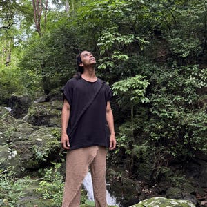 May include: A person wearing a black short-sleeved top with a diagonal strap and tan pants. The person is standing in a lush, green forest with a waterfall in the background. The clothing appears to be made of natural fibers.