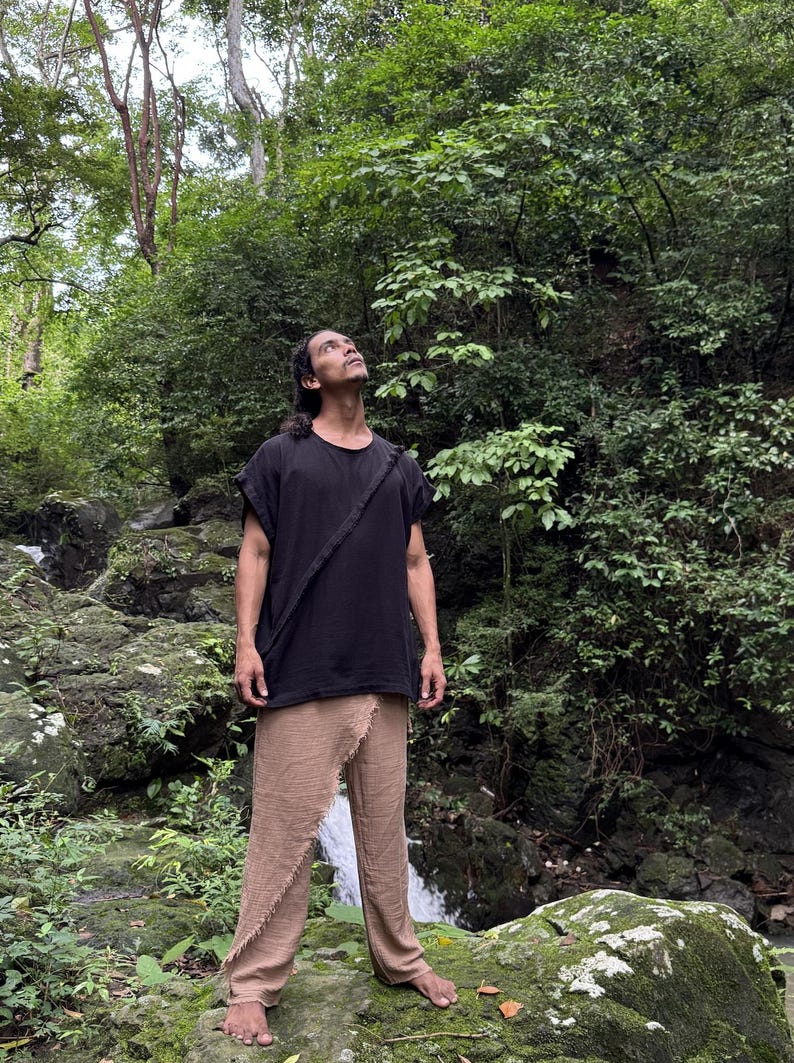 May include: A person wearing a black short-sleeved top with a diagonal strap and tan pants. The person is standing in a lush, green forest with a waterfall in the background. The clothing appears to be made of natural fibers.