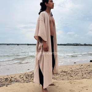 Pu&ograve; includere: Un abito a kimono beige con apertura frontale, maniche ampie e aspetto strutturato. L'abito &egrave; indossato sopra pantaloni neri. L'immagine include il testo "www.heartearthcraft.com" in basso. La persona &egrave; in piedi su una spiaggia sabbiosa.