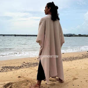 Pu&ograve; includere: Un lungo cardigan aperto beige chiaro con maniche ampie. Il cardigan &egrave; indossato sopra pantaloni neri. L'immagine &egrave; scattata su una spiaggia sabbiosa con l'oceano sullo sfondo. L'indirizzo del sito web "WWW.HEARTEARTHCRAFT.COM" &egrave; visibile.