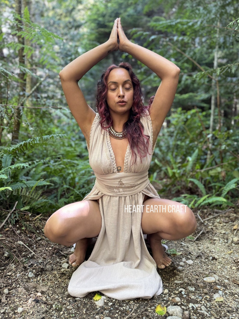 May include: A woman wearing a beige maxi dress with a deep V-neckline and a slit up the front. She is sitting in a squat position with her hands raised in a prayer position. The dress has a brown beaded necklace and the text "HEART EARTH CRAFT" is visible on the bottom of the dress.