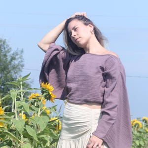 May include: A woman wearing a purple off-the-shoulder top with long sleeves and a white skirt with a side slit. She is standing in a field of sunflowers.