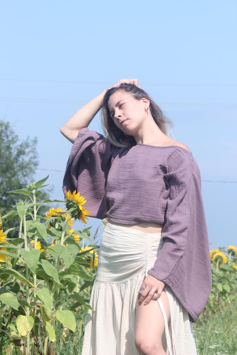 May include: A woman wearing a purple off-the-shoulder top with long sleeves and a white skirt with a side slit. She is standing in a field of sunflowers.
