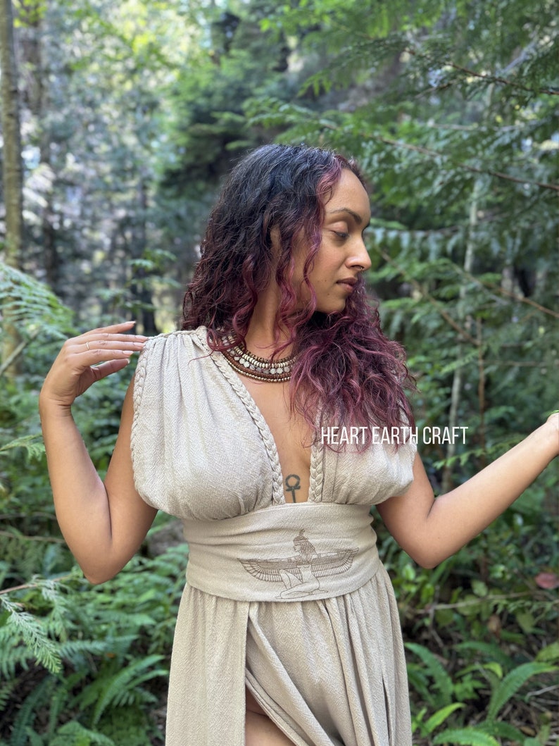 May include: A woman wearing a beige linen dress with a deep V-neckline and a decorative belt featuring an embroidered winged figure. The dress has a slit in the front and the woman is standing in a forest setting. The text "HEART EARTH CRAFT" is visible on the dress.