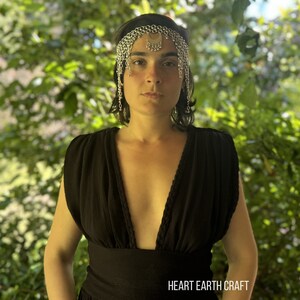 May include: A woman wearing a black sleeveless dress with a plunging neckline and a silver chain headpiece. The text "HEART EARTH CRAFT" is visible at the bottom of the image.