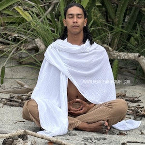 Pu&ograve; includere: Una persona seduta a gambe incrociate sul terreno, con un scialle di cotone bianco e pantaloni marroni. Ha gli occhi chiusi e le mani in posizione di meditazione. Lo sfondo &egrave; un'immagine sfocata di piante verdi.
