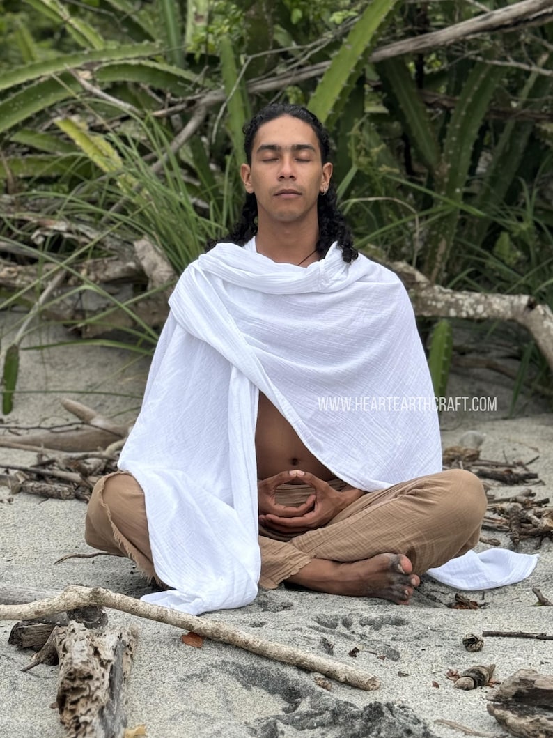 Pu&ograve; includere: Una persona seduta a gambe incrociate sul terreno, con un scialle di cotone bianco e pantaloni marroni. Ha gli occhi chiusi e le mani in posizione di meditazione. Lo sfondo &egrave; un'immagine sfocata di piante verdi.
