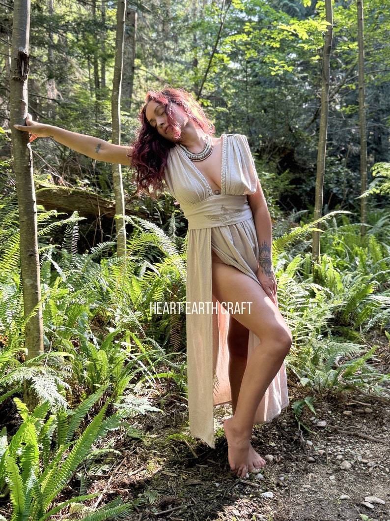 May include: A woman wearing a long, light brown linen dress with a deep V-neckline and a cinched waist. The dress has a slit up the side and is flowing in the breeze. She is standing in a forest setting with ferns and trees surrounding her. The text "HEART EARTH CRAFT" is visible on the dress.
