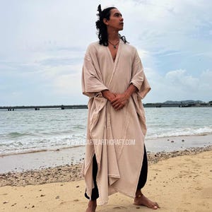 Pu&ograve; includere: Una veste beige con apertura frontale, maniche ampie e orlo spaccato. La veste &egrave; indossata sopra pantaloni neri. L'immagine &egrave; scattata su una spiaggia sabbiosa con l'oceano sullo sfondo. Il sito web www.heartearthcraft.com &egrave; visibile.