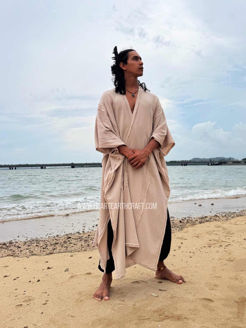 Pu&ograve; includere: Una veste beige con apertura frontale, maniche ampie e orlo spaccato. La veste &egrave; indossata sopra pantaloni neri. L'immagine &egrave; scattata su una spiaggia sabbiosa con l'oceano sullo sfondo. Il sito web www.heartearthcraft.com &egrave; visibile.