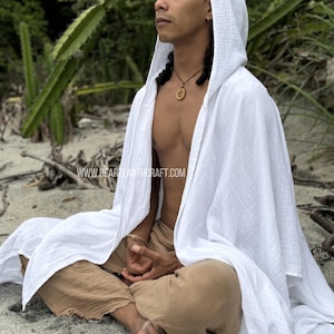 Pu&ograve; includere: Una persona indossa una tunica bianca con cappuccio e siede a gambe incrociate su una spiaggia di sabbia. La tunica &egrave; realizzata in un tessuto leggero e traspirante e ha una vestibilit&agrave; rilassata. La persona indossa pantaloni marroni e ha una collana con un ciondolo di legno.
