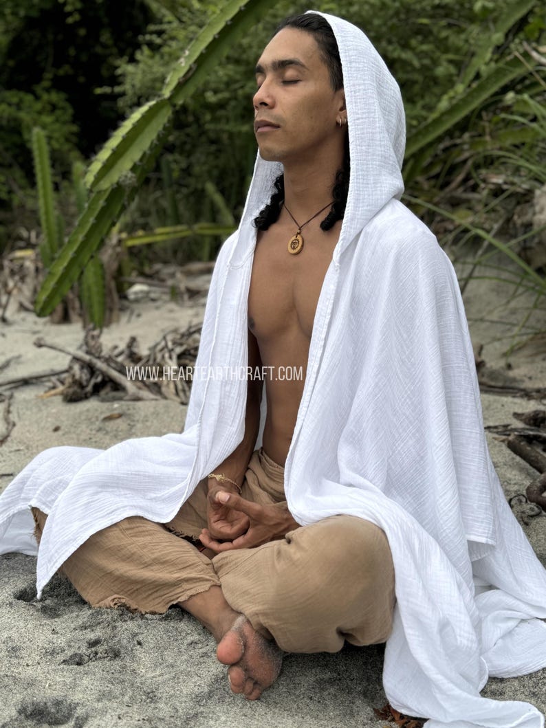 Pu&ograve; includere: Una persona indossa una tunica bianca con cappuccio e siede a gambe incrociate su una spiaggia di sabbia. La tunica &egrave; realizzata in un tessuto leggero e traspirante e ha una vestibilit&agrave; rilassata. La persona indossa pantaloni marroni e ha una collana con un ciondolo di legno.