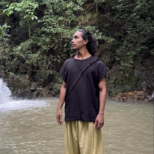 May include: A person wearing a black short-sleeved shirt with a strap across the chest and light yellow pants. The person is standing in front of a waterfall and a body of water, surrounded by lush green foliage.