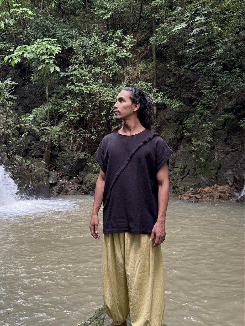 May include: A person wearing a black short-sleeved shirt with a strap across the chest and light yellow pants. The person is standing in front of a waterfall and a body of water, surrounded by lush green foliage.