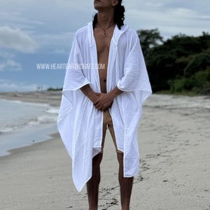 Pu&ograve; includere: Una lunga kaftan di cotone bianco con cappuccio e un design fluido. La kaftan &egrave; aperta sul davanti e ha una vestibilit&agrave; rilassata. Il modello &egrave; in piedi su una spiaggia di sabbia con l'oceano sullo sfondo. L'indirizzo del sito web www.hearthcraft.com &egrave; visibile nell'angolo in basso a sinistra dell'immagine.