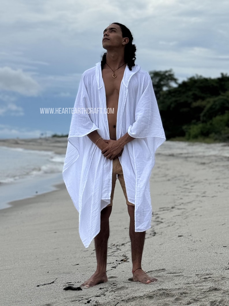 Pu&ograve; includere: Una lunga kaftan di cotone bianco con cappuccio e un design fluido. La kaftan &egrave; aperta sul davanti e ha una vestibilit&agrave; rilassata. Il modello &egrave; in piedi su una spiaggia di sabbia con l'oceano sullo sfondo. L'indirizzo del sito web www.hearthcraft.com &egrave; visibile nell'angolo in basso a sinistra dell'immagine.