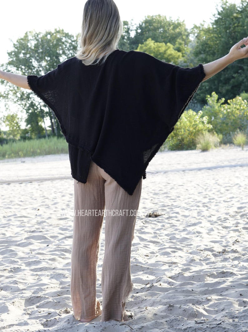 May include: A black, loose-fitting, short-sleeved top with a raw edge hem. The top is worn with light brown, wide-leg pants. The model is standing on a sandy beach.