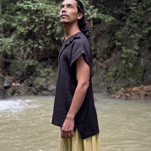May include: A person wearing a black short-sleeved top with a textured neckline and a yellow skirt. The person is standing near a body of water with a lush green background.