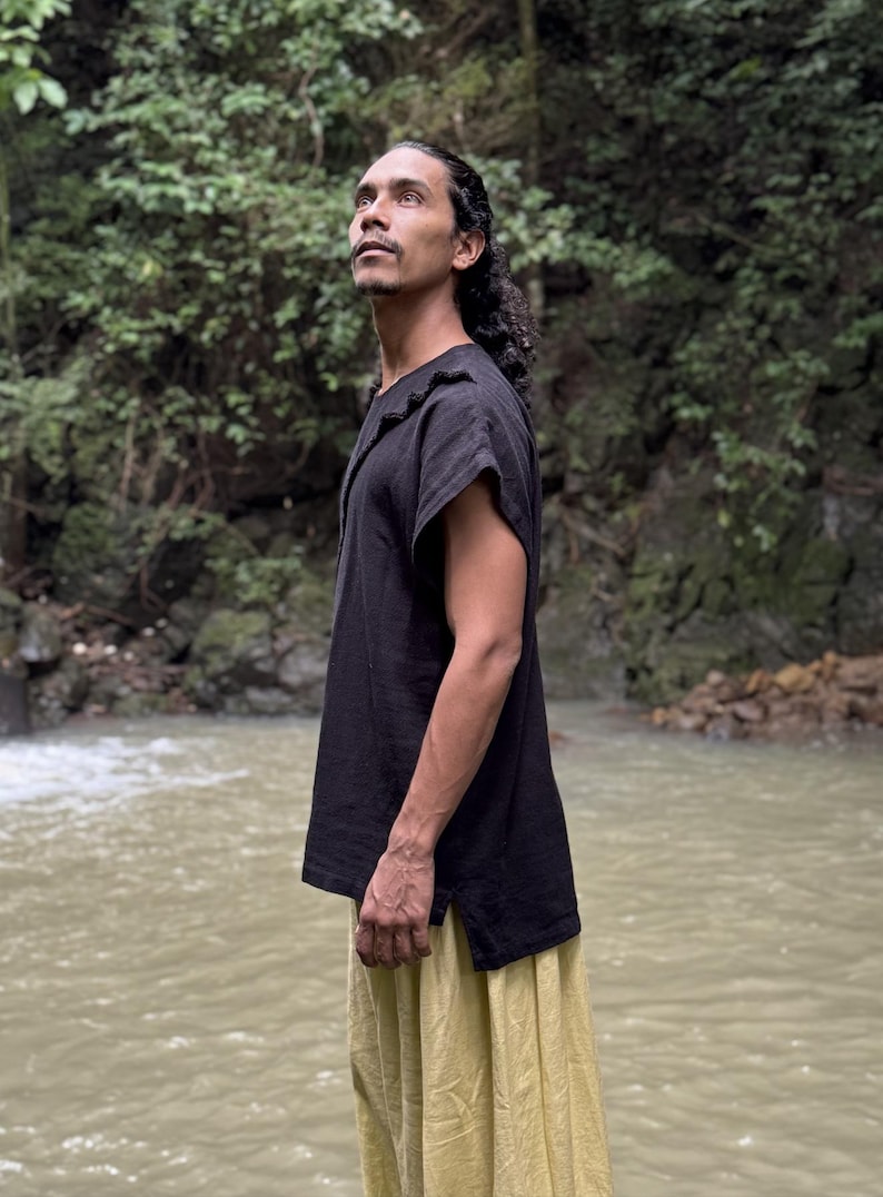 May include: A person wearing a black short-sleeved top with a textured neckline and a yellow skirt. The person is standing near a body of water with a lush green background.