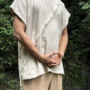 May include: A beige, short-sleeved shirt with a diagonal strap detail and matching beige pants. The shirt has a textured appearance and a round neckline. The pants are loose-fitting with a vertical striped pattern. The outfit is made of a lightweight fabric.