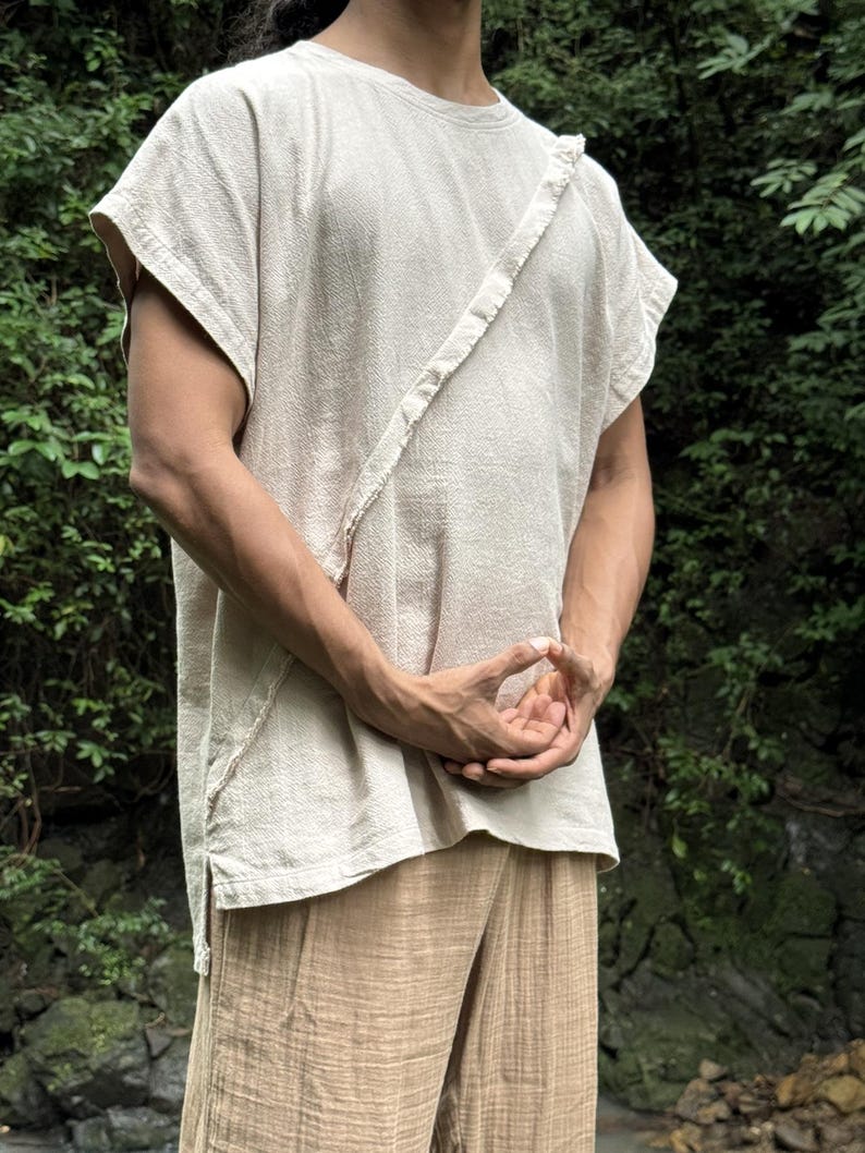 May include: A beige, short-sleeved shirt with a diagonal strap detail and matching beige pants. The shirt has a textured appearance and a round neckline. The pants are loose-fitting with a vertical striped pattern. The outfit is made of a lightweight fabric.