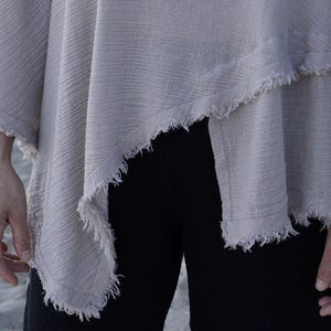 May include: A light gray, loose-fitting,  cotton top with a frayed hem. The top is draped over one shoulder and has a relaxed, casual style.