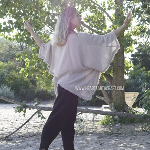 May include: A woman wearing a light beige, loose-fitting,  cotton top with a frayed hem. She is standing on a sandy beach with trees in the background. The top has a relaxed, flowy silhouette and is perfect for a casual, bohemian look.  The text "www.hearthcraft.com" is visible on the bottom of the image.