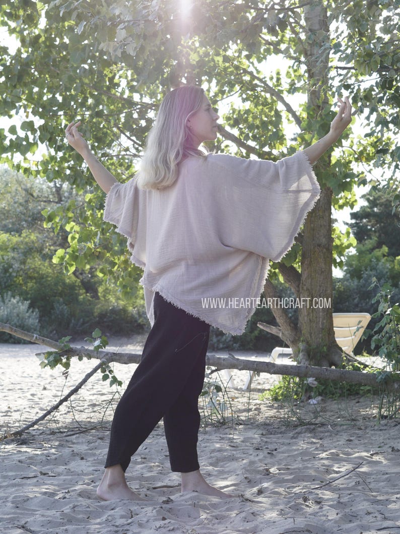 May include: A woman wearing a light beige, loose-fitting,  cotton top with a frayed hem. She is standing on a sandy beach with trees in the background. The top has a relaxed, flowy silhouette and is perfect for a casual, bohemian look.  The text "www.hearthcraft.com" is visible on the bottom of the image.