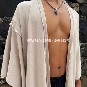 Pu&ograve; includere: Un accappatoio beige in stile kimono con apertura frontale e bordi sfilacciati. L'accappatoio &egrave; indossato sopra pantaloni neri e una collana marrone con un ciondolo turchese. L'indirizzo del sito web "WWW.HEARTEARTHCRAFT.COM" &egrave; visibile.