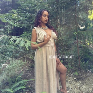 May include: A woman wearing a long, light brown linen dress with a deep V-neckline and a slit up the side. She is standing in a forest setting with green foliage and trees in the background. The dress has a simple design and is made of a natural fabric. The text "HEART EARTH CRAFT" is visible in the lower right corner of the image.