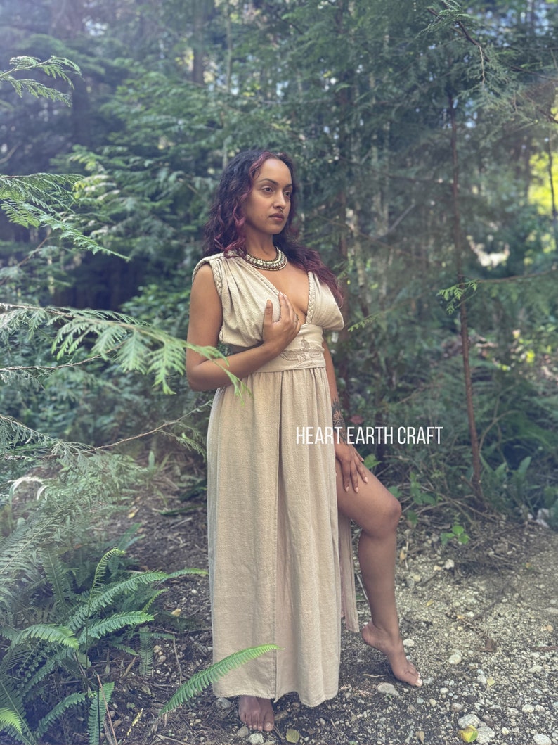 May include: A woman wearing a long, light brown linen dress with a deep V-neckline and a slit up the side. She is standing in a forest setting with green foliage and trees in the background. The dress has a simple design and is made of a natural fabric. The text "HEART EARTH CRAFT" is visible in the lower right corner of the image.
