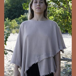 May include: A light beige, loose-fitting, asymmetrical top with frayed edges. The top is made of a soft, textured fabric and has a relaxed fit. The model is wearing the top with black pants. The text "www.hearthcraft.com" is visible at the bottom of the image.