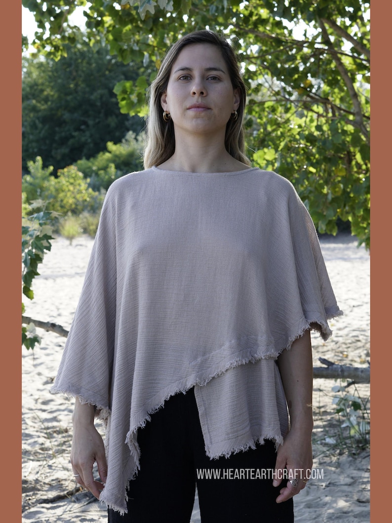 May include: A light beige, loose-fitting, asymmetrical top with frayed edges. The top is made of a soft, textured fabric and has a relaxed fit. The model is wearing the top with black pants. The text "www.hearthcraft.com" is visible at the bottom of the image.