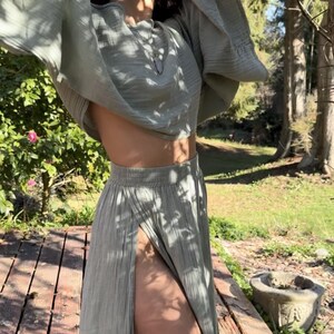 May include: A sage green two-piece set consisting of a cropped top with wide, flared sleeves and a matching high-waisted maxi skirt featuring a high front slit. The fabric has a textured, crinkled appearance and a soft, matte finish throughout.