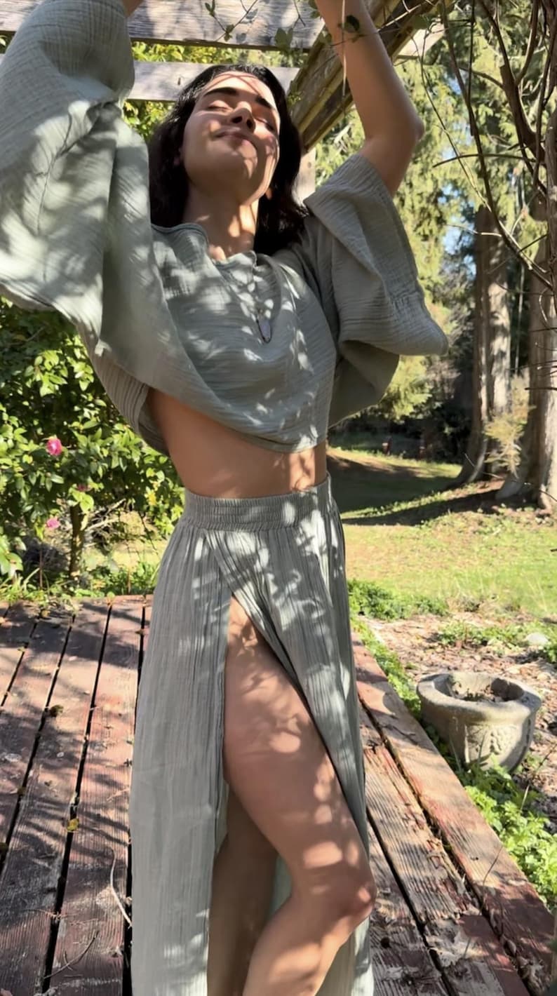 May include: A sage green two-piece set consisting of a cropped top with wide, flared sleeves and a matching high-waisted maxi skirt featuring a high front slit. The fabric has a textured, crinkled appearance and a soft, matte finish throughout.