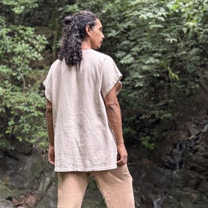 May include: A beige linen tunic and matching pants. The short-sleeved tunic has a relaxed fit, and the pants have a loose, textured appearance. The outfit is worn by a person with long, dark hair, standing in a natural outdoor setting.