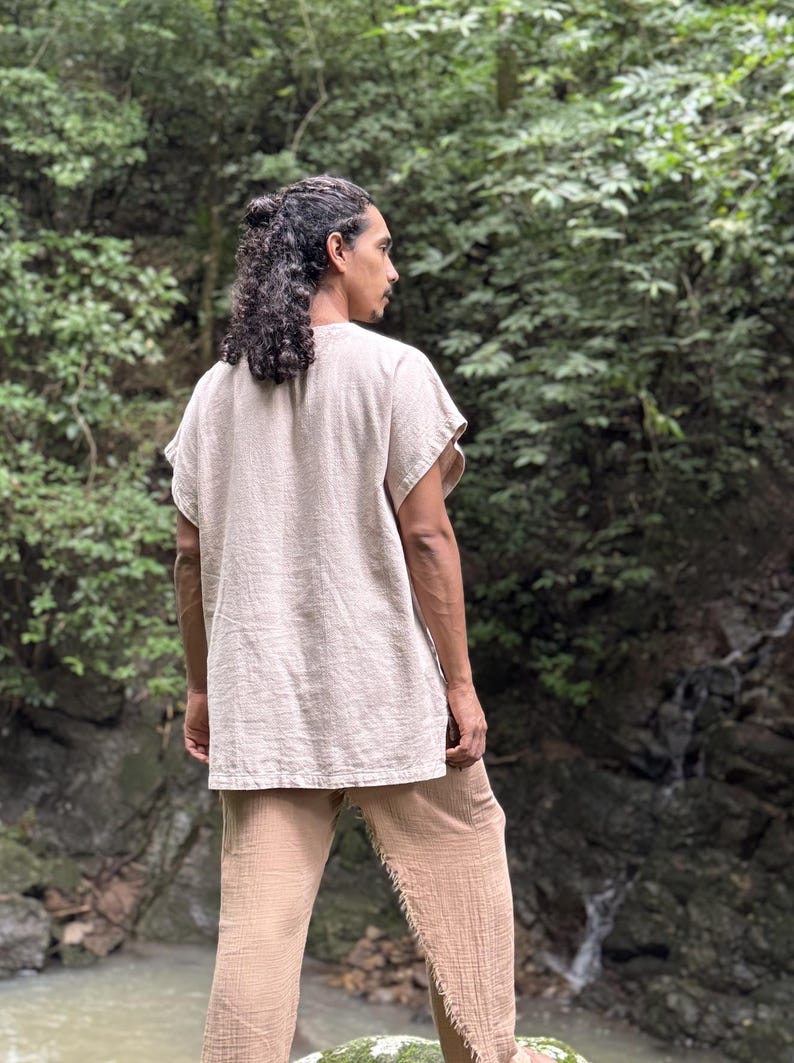 May include: A beige linen tunic and matching pants. The short-sleeved tunic has a relaxed fit, and the pants have a loose, textured appearance. The outfit is worn by a person with long, dark hair, standing in a natural outdoor setting.