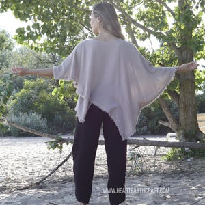 May include: A woman wearing a light brown, loose-fitting, asymmetrical top with a frayed hem and black pants. The top has a wide, flowing silhouette. The woman is standing on a sandy beach with trees in the background. The text "www.hearthcraft.com" is visible at the bottom of the image.